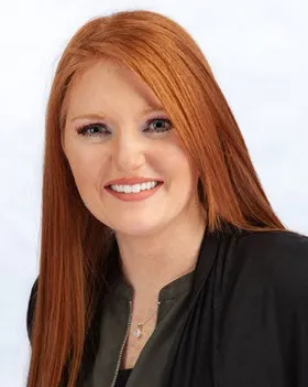 A woman with long straight red hair smiles at the camera. She is wearing a dark top and a black jacket, with a simple necklace. The background is light and plain.