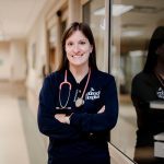 A smiling healthcare professional stands in a hospital hallway, wearing a navy shirt with Anderson Hospital on it and a stethoscope around her neck. Her arms are crossed and she faces the camera confidently.