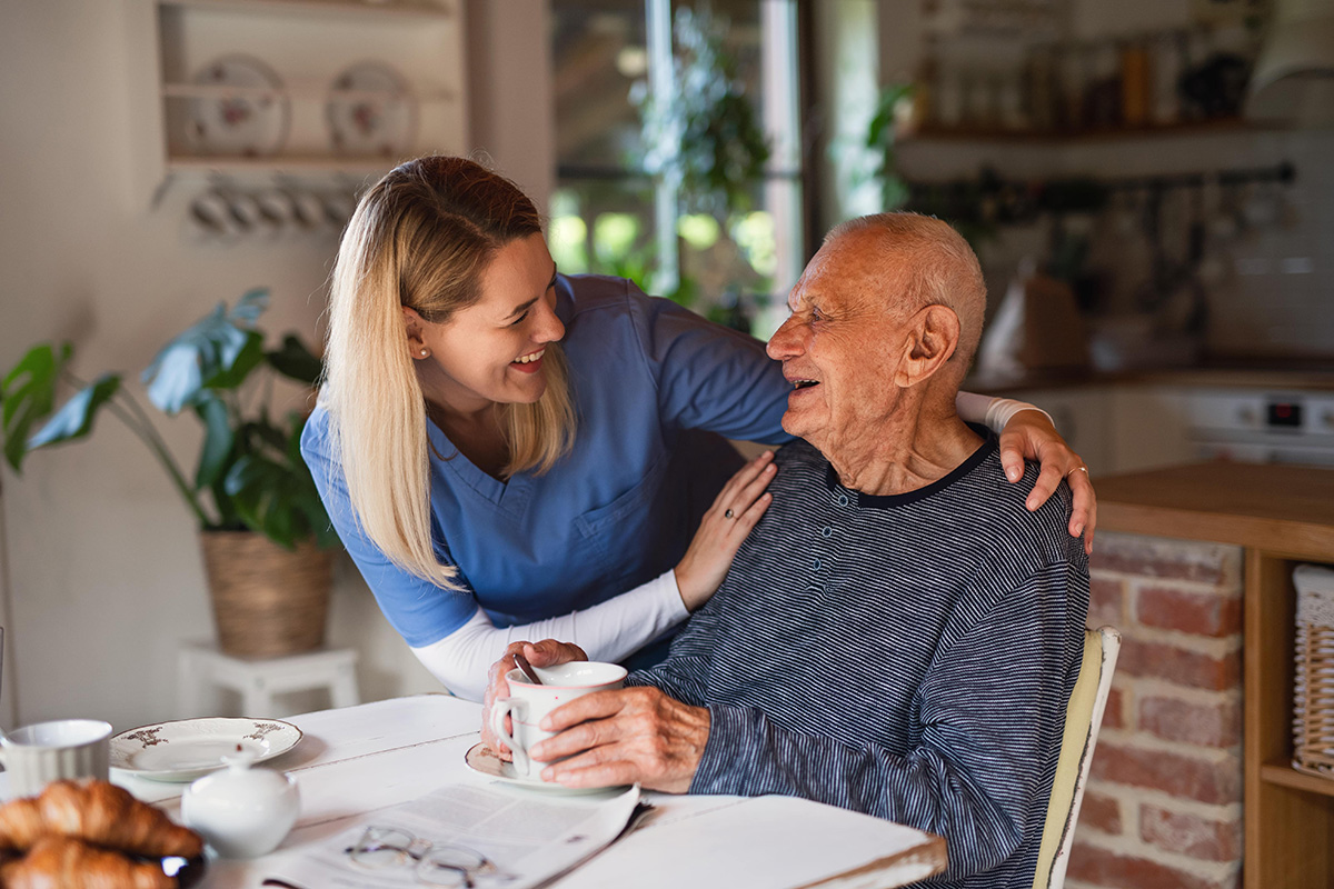 A smiling caregiver in blue scrubs stands beside an elderly man sitting at a kitchen table, both laughing and enjoying a warm moment together. The man holds a cup, and sunlight streams into the cozy room.