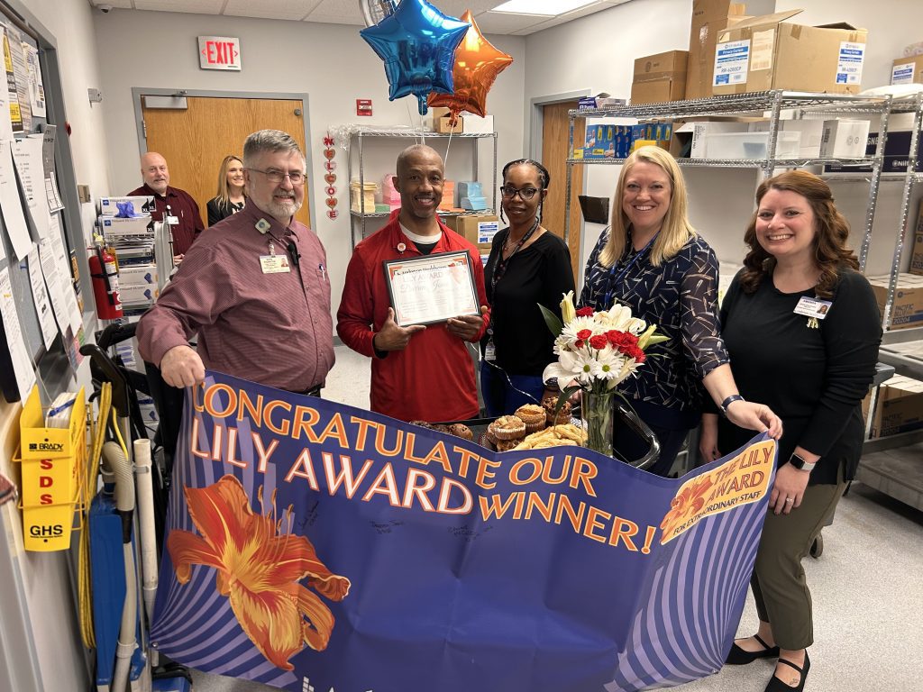 Five people stand in a medical supply room, smiling and holding a certificate. A celebratory banner reads “CONGRATULATE OUR LILLY AWARD WINNER!” with flowers, balloons, and cupcakes on the table.