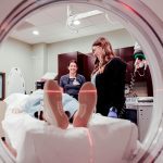 A patient lies on a hospital bed entering a CT scanner, viewed from inside the machine, while two healthcare workers stand nearby, one smiling and the other preparing equipment.