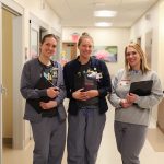 Three smiling healthcare workers with clipboards stand in a well-lit hospital hallway, wearing scrubs and ID badges. They appear friendly and approachable, ready to assist patients.