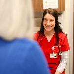 A smiling healthcare professional in red scrubs speaks with a patient, who is seen from behind, in a medical office setting. The healthcare worker has a stethoscope and name badge.