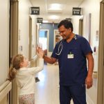 A doctor in blue scrubs gives a high-five to a smiling young girl in the hallway of the world’s best hospital, with exam room signs visible on the doors.