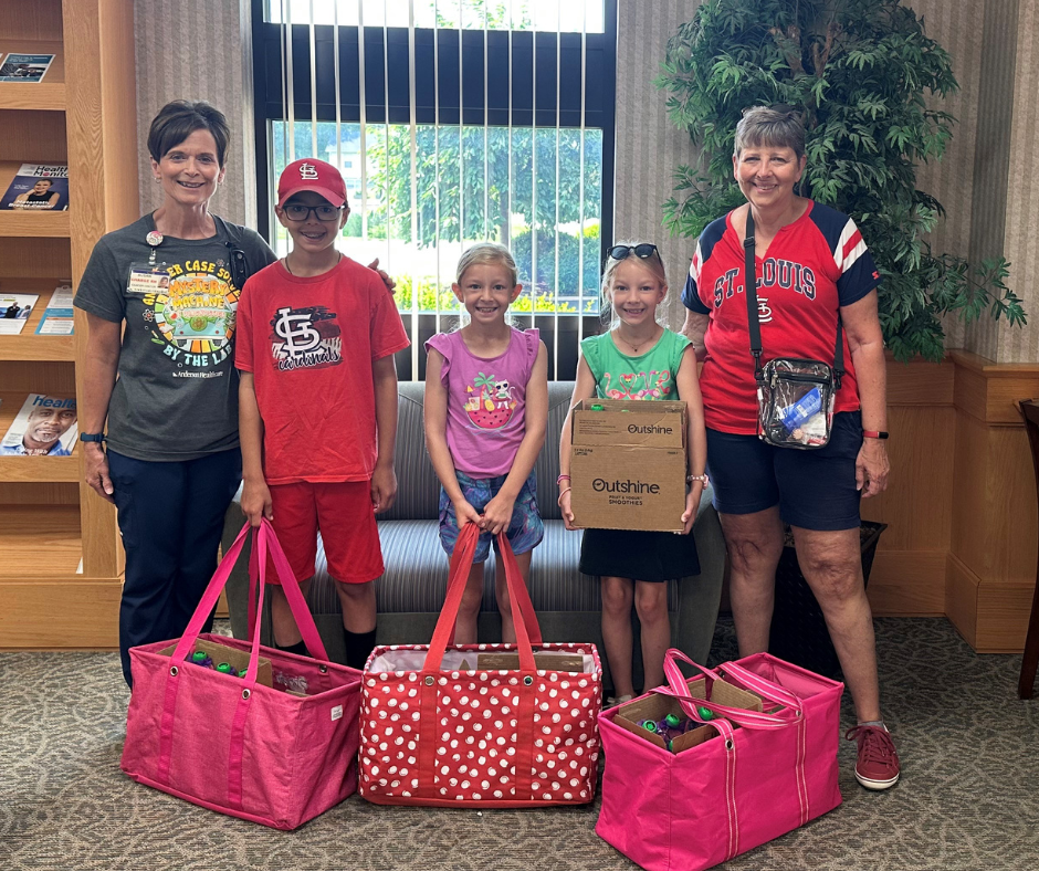 Four smiling children and two women stand indoors with bags and a box of supplies. The children are holding a box and standing behind pink and polka dot bags. Bookshelves and a large window are in the background.