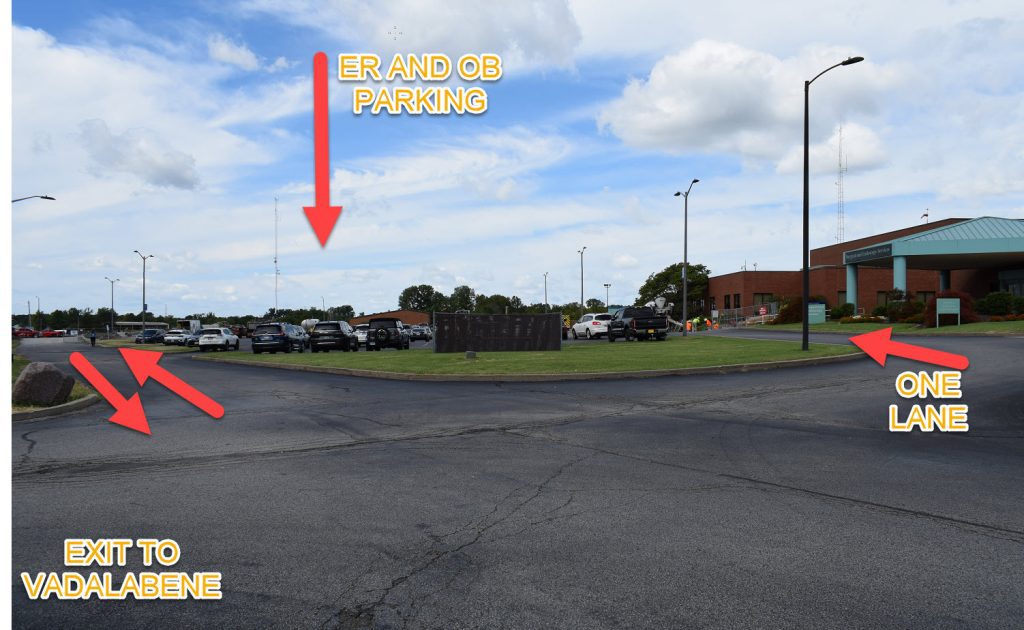 Hospital parking lot scene with labeled arrows: ER AND OB PARKING points to a lot, EXIT TO VADALABENE points left, and ONE LANE points right toward a single-lane drive near a brick building.