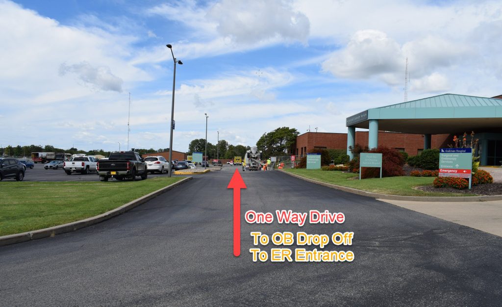 A hospital driveway with a large red arrow and text that reads: One Way Drive To OB Drop Off To ER Entrance pointing toward the entrance of the building under a partly cloudy sky.
