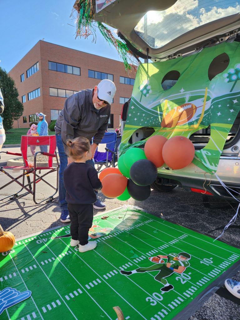 A man and a young child stand near a decorated car trunk with a football theme. The trunk has brown and black balloons and a game backdrop. They are on a mat designed like a football field in a parking lot.