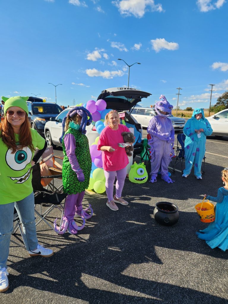 A group of people in colorful Monsters, Inc. costumes stand by a car decorated with balloons in a parking lot on a sunny day. One person holds a small dog, and Halloween buckets are on the ground.