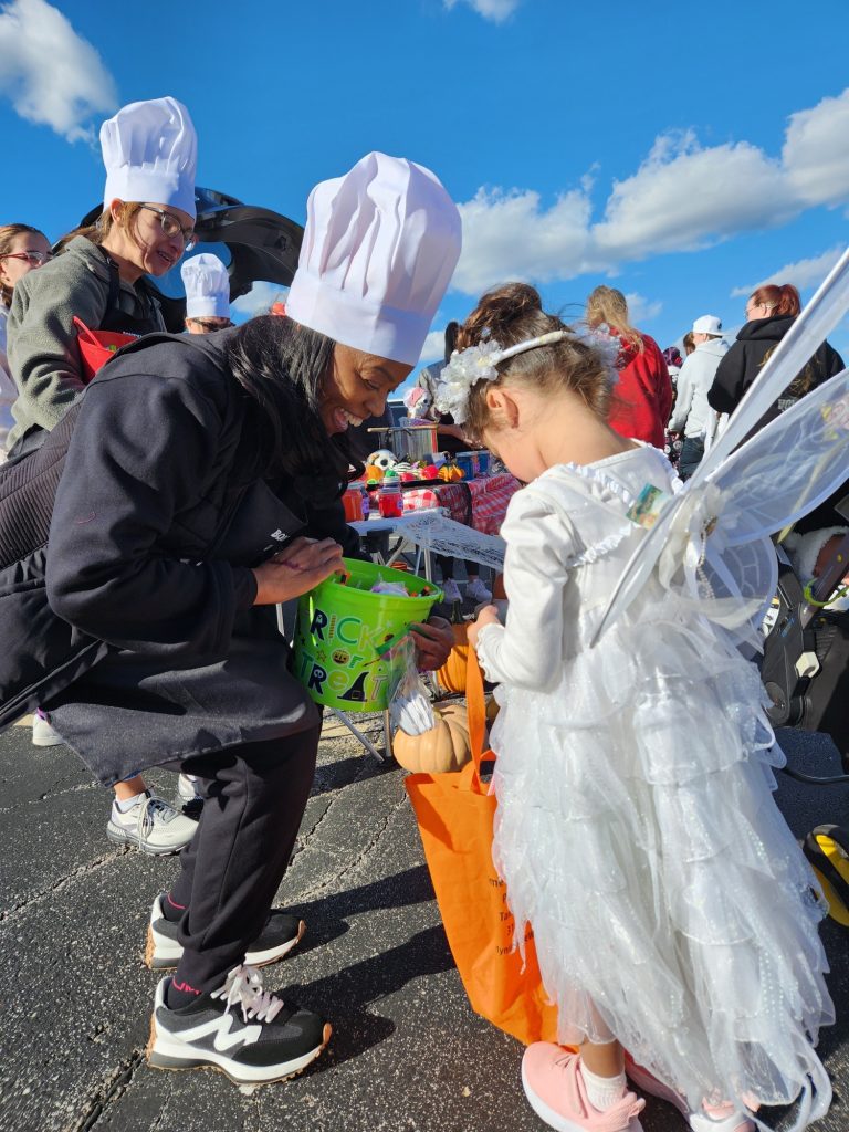 A woman in a chef hat kneels beside a young girl dressed as a fairy with wings and a white dress. They are outdoors at a festive event, collecting treats. Other people and a table of items are visible in the background.