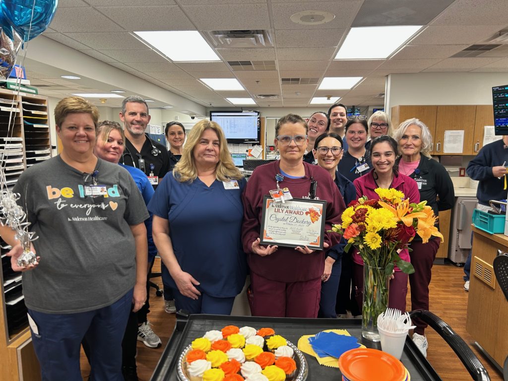 A group of healthcare workers poses in a hospital unit. One person in the center holds a DAISY Award certificate. There are balloons, autumn-themed cupcakes, cookies, and a vase of flowers on the counter in front of them.