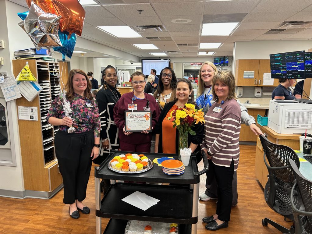 A group of eight smiling healthcare workers stand around a cart with cupcakes, flowers, and plates in a hospital setting. One person in the center holds a certificate, and balloons are visible on the left.