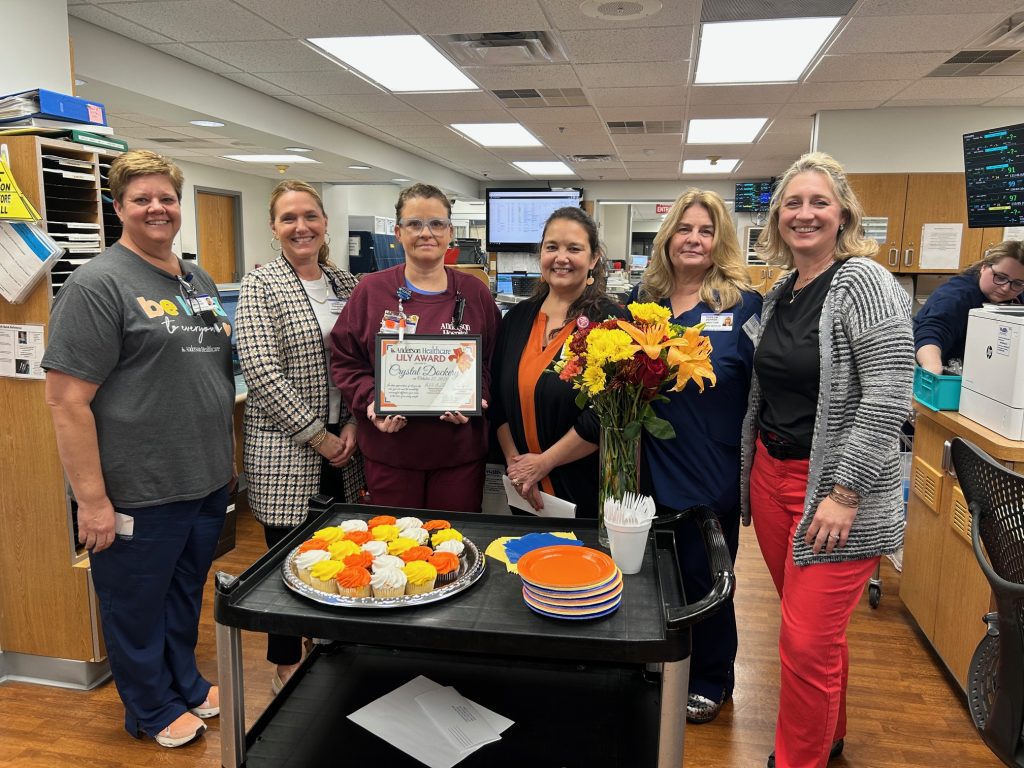 Six women stand smiling around a cart with cupcakes, plates, and a bouquet of flowers in an office or hospital setting. One woman holds a certificate. Computer monitors and office supplies are visible in the background.