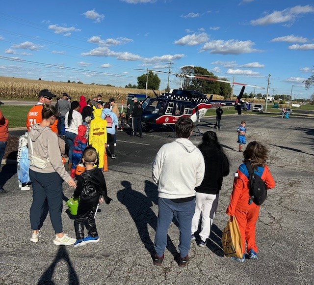 A group of people, including children in costumes, stand in a parking lot on a sunny day, watching a red and black helicopter parked nearby. Fields and a clear sky with scattered clouds are in the background.