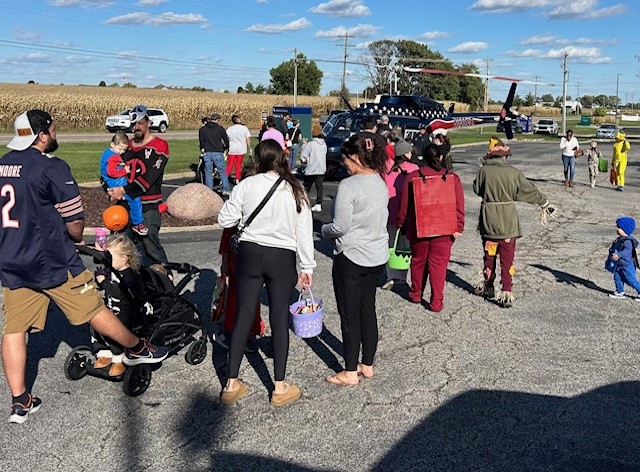 A group of people, some with children and strollers, gather outdoors near a parked helicopter on a sunny day. Some children hold baskets, possibly for trick-or-treating. Cornfields and cars are visible in the background.