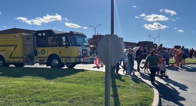 A yellow fire truck is parked near a group of people, some in costumes, walking on a sunny day. The scene appears to be part of an outdoor community event near a building.