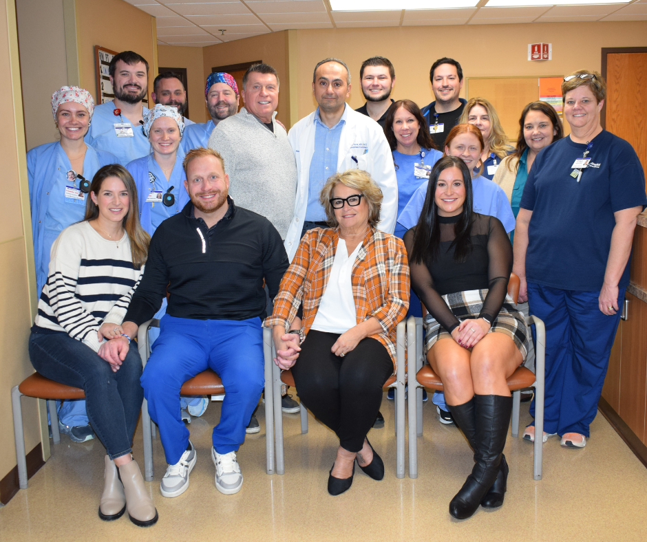 A group of people, including medical staff in scrubs and others in casual clothing, pose together in a hallway. Four people are seated in front while the rest stand behind them, all smiling at the camera.