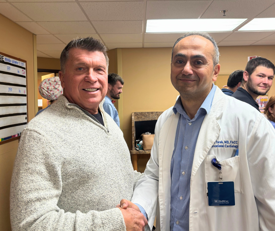 Two men are smiling and shaking hands in a medical office. One man is wearing a white coat with a name badge, indicating he is a doctor. Several other people are visible in the background.