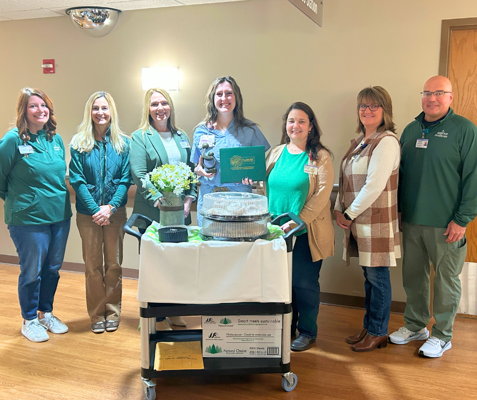Seven people stand smiling behind a cart with food trays, a bouquet of daisies, and a certificate in a hallway. One woman in the center holds a green folder and trophy. Everyone is dressed in business casual or uniforms.