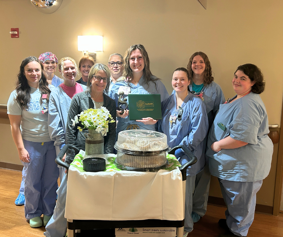 A group of smiling people in scrubs and casual clothes gather around a cart with flowers and covered trays in a well-lit room. One person in the center holds a Daisy award certificate, celebrating their caring achievements.
