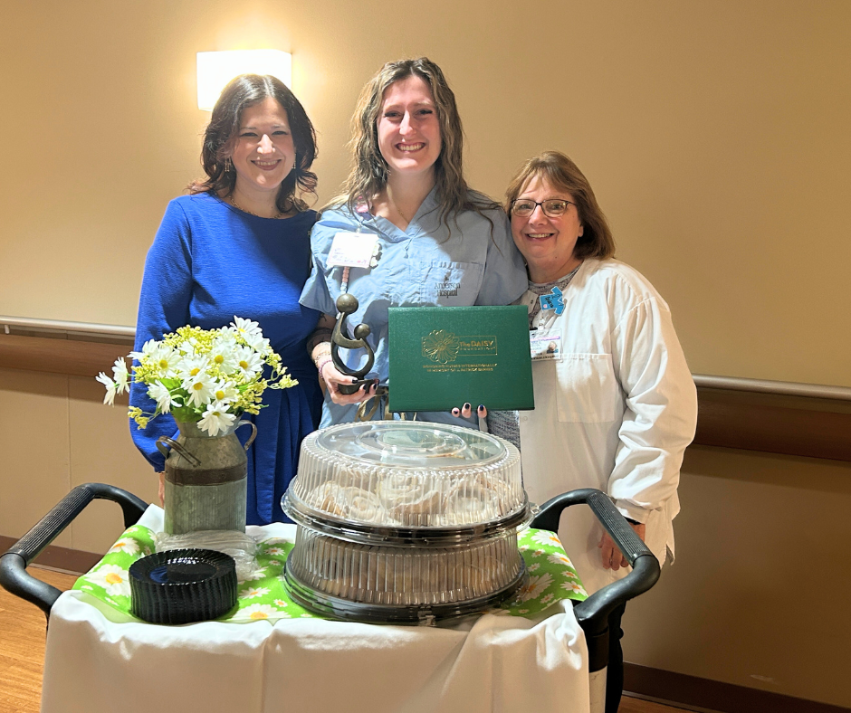 Three women stand smiling behind a cart with flowers and a tray of pastries. The woman in the center, wearing scrubs, holds a Daisy award and certificate. The other women are dressed in blue and white, indoors in a well-lit hallway.