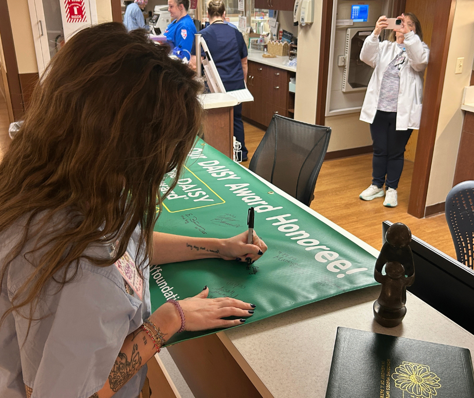 A woman signs a green Daisy Award Honoree banner at a hospital nurses station while a nurse in the background takes a photo on her phone. Other staff members celebrate the special Daisy recognition moment.