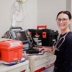 A healthcare professional with glasses and a stethoscope smiles while standing next to medical equipment and a crash cart in a hospital or clinical setting.