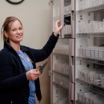 A healthcare worker in scrubs and a navy jacket smiles while organizing or retrieving medication packets from a storage cabinet in a medical facility.