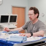 A man wearing glasses and an Anderson Hospital t-shirt prepares medical supplies on a tray in a clinical room with medical monitors and equipment in the background.