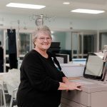 A smiling woman with short gray hair and glasses stands beside a medical machine in a hospital or clinic setting, wearing a black top. Medical equipment and monitors are visible in the background.