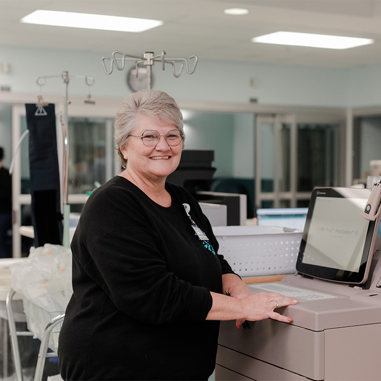 A smiling woman with short gray hair and glasses stands beside a medical machine in a hospital or clinic setting, wearing a black top. Medical equipment and monitors are visible in the background.