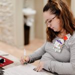 A woman with glasses and long hair writes on a form at a desk. She wears a gray shirt, name badge, and a cat-shaped badge reel. A red mouse pad and computer mouse are nearby. The background is softly blurred.