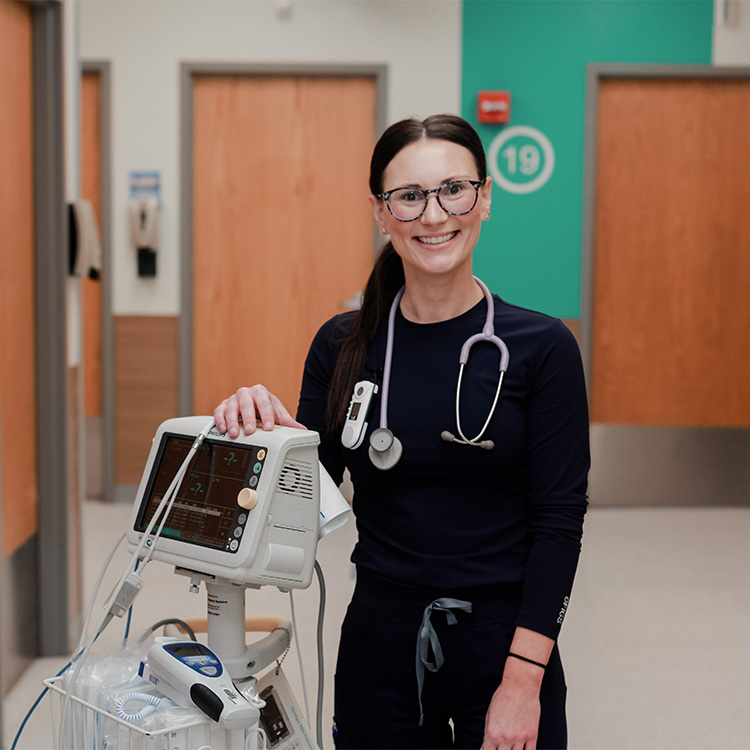 A smiling healthcare professional with a stethoscope stands beside medical equipment in a hospital hallway, wearing navy scrubs and glasses.
