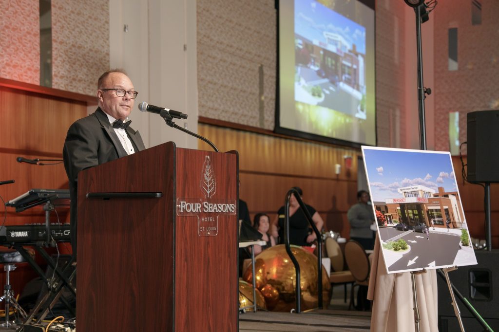 A man in a tuxedo speaks at a podium labeled Four Seasons Hotel St. Louis in a banquet hall; a large poster and projected image show an architectural rendering of a grocery store.