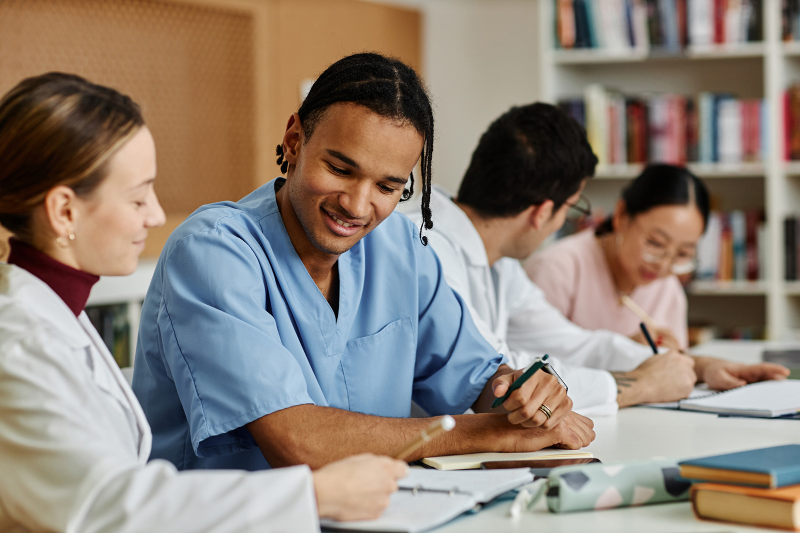 Four medical students, two men and two women, sit at a table in a library, studying and writing in notebooks. One man in blue scrubs smiles while talking with a woman in a white coat. Bookshelves are in the background.