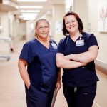 Two smiling healthcare workers in navy scrubs stand in a hospital hallway, with one holding a clipboard. They both wear name badges and stand in front of light-colored walls decorated with artwork.