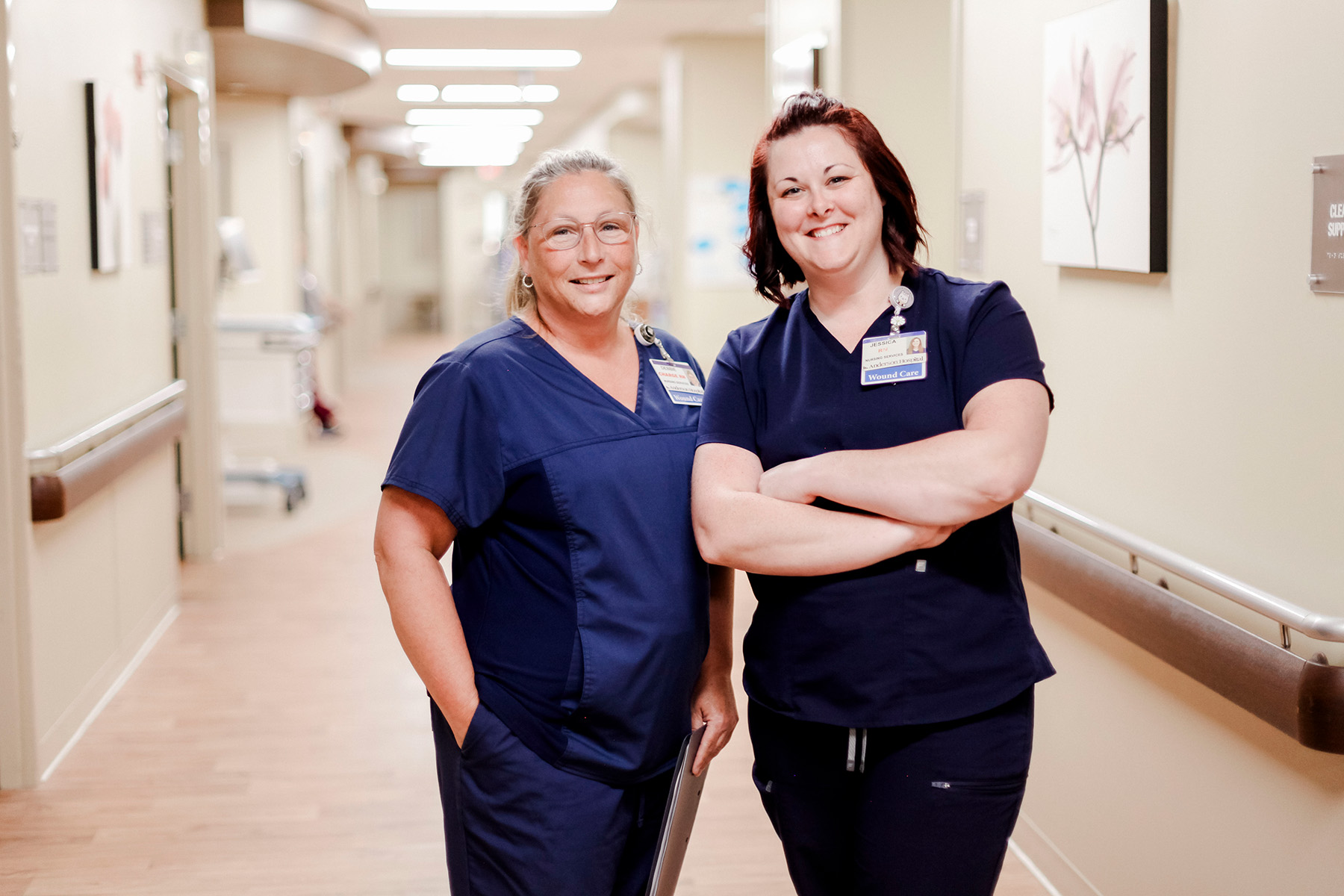 Two smiling healthcare workers in navy scrubs stand in a hospital hallway, with one holding a clipboard. They both wear name badges and stand in front of light-colored walls decorated with artwork.