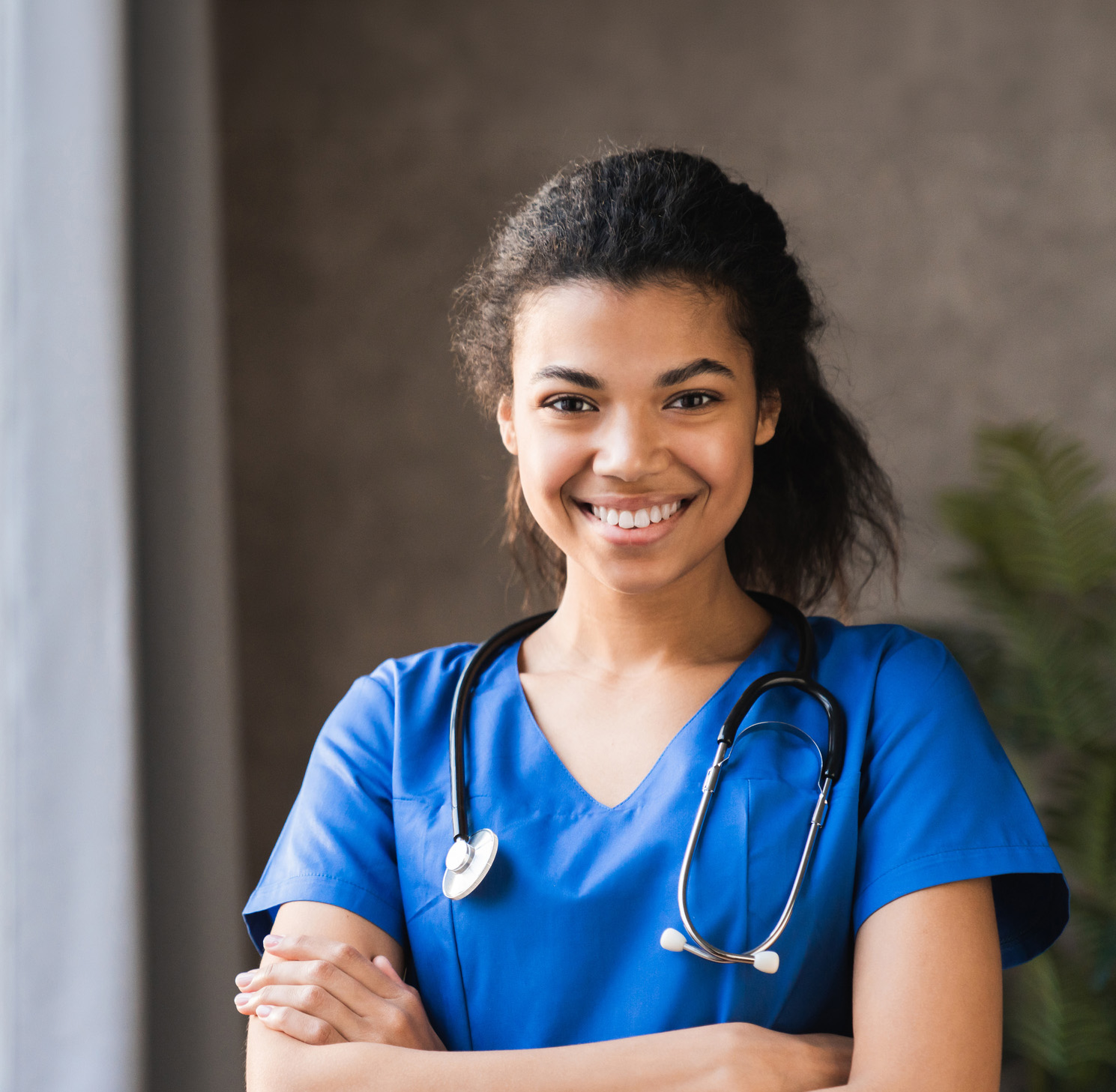 A smiling woman in blue medical scrubs with a stethoscope around her neck stands with arms crossed in a brightly lit room.