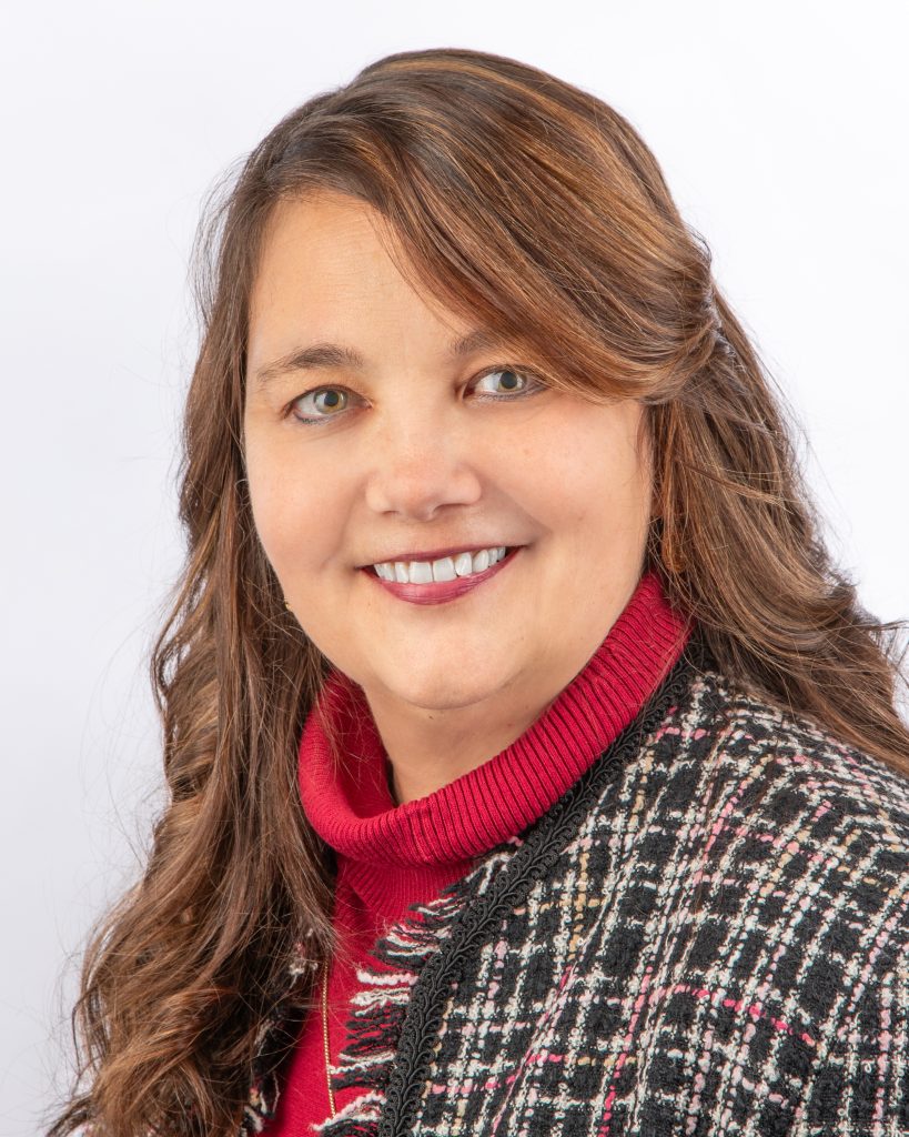 A woman with long, wavy brown hair smiles at the camera. She is wearing a red turtleneck sweater under a black and white plaid jacket, and is posed in front of a plain white background.