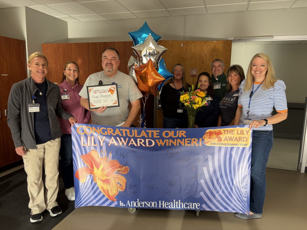 A group of smiling hospital staff stands indoors holding a blue and gold banner that says CONGRATULATE OUR LILY AWARD WINNER! Anderson Healthcare. One man holds a certificate and balloons; a woman holds flowers.