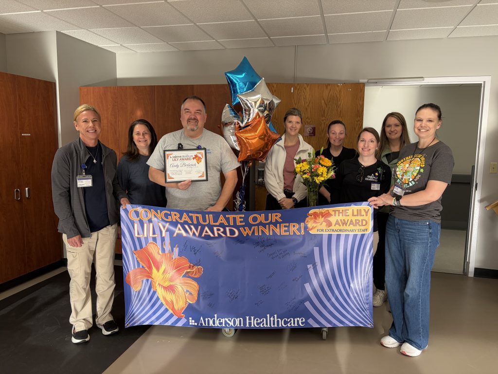 A group of people smile while holding a Lily Award Winner banner, balloons, flowers, and a certificate inside a room at Anderson Healthcare.