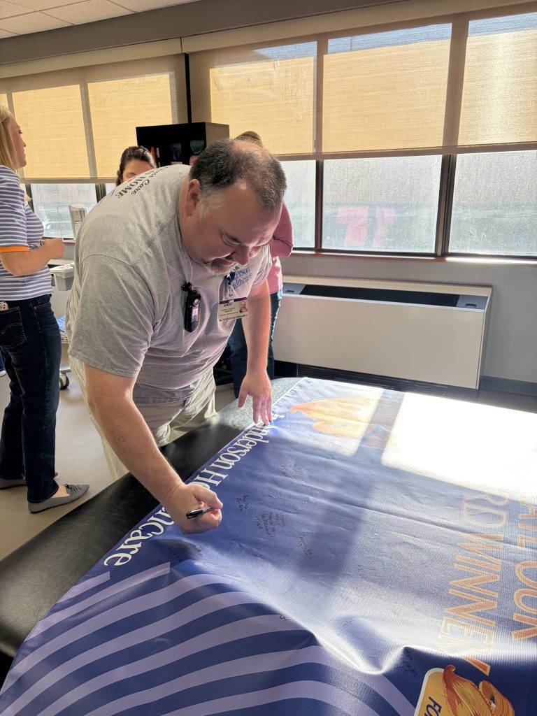 A man in a gray shirt leans over a table and writes on a large, blue banner with a marker while other people stand nearby in a well-lit room with large windows.