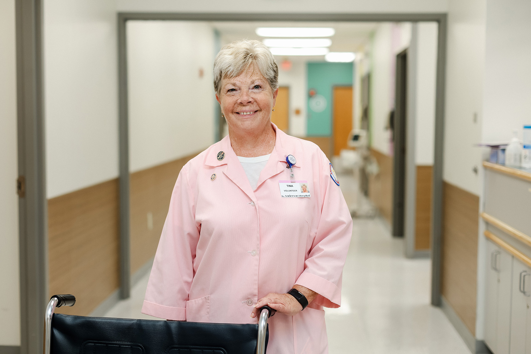An older woman in a pink medical uniform stands smiling behind a wheelchair in a bright, clean hospital hallway.