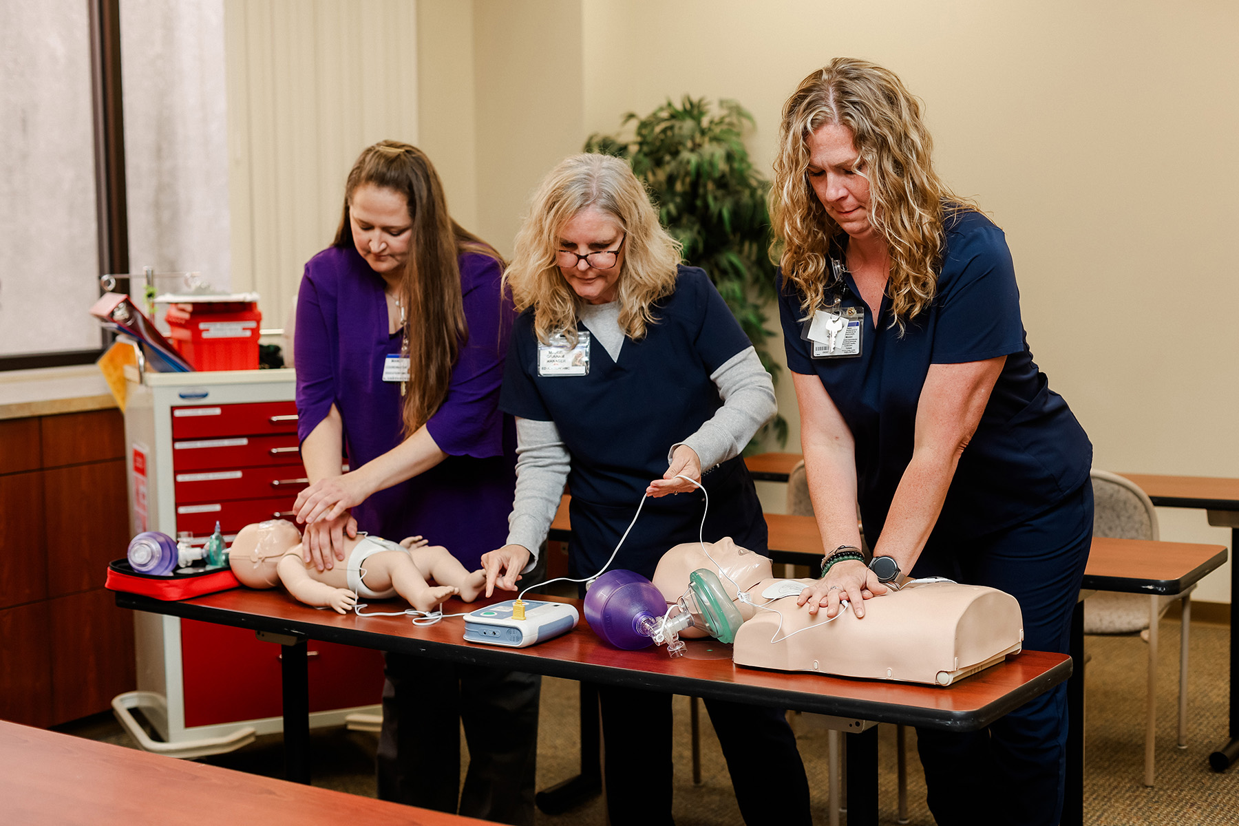 Three women in medical scrubs practice CPR on infant and adult mannequins using medical equipment in a classroom setting, focused on a life-saving training exercise.