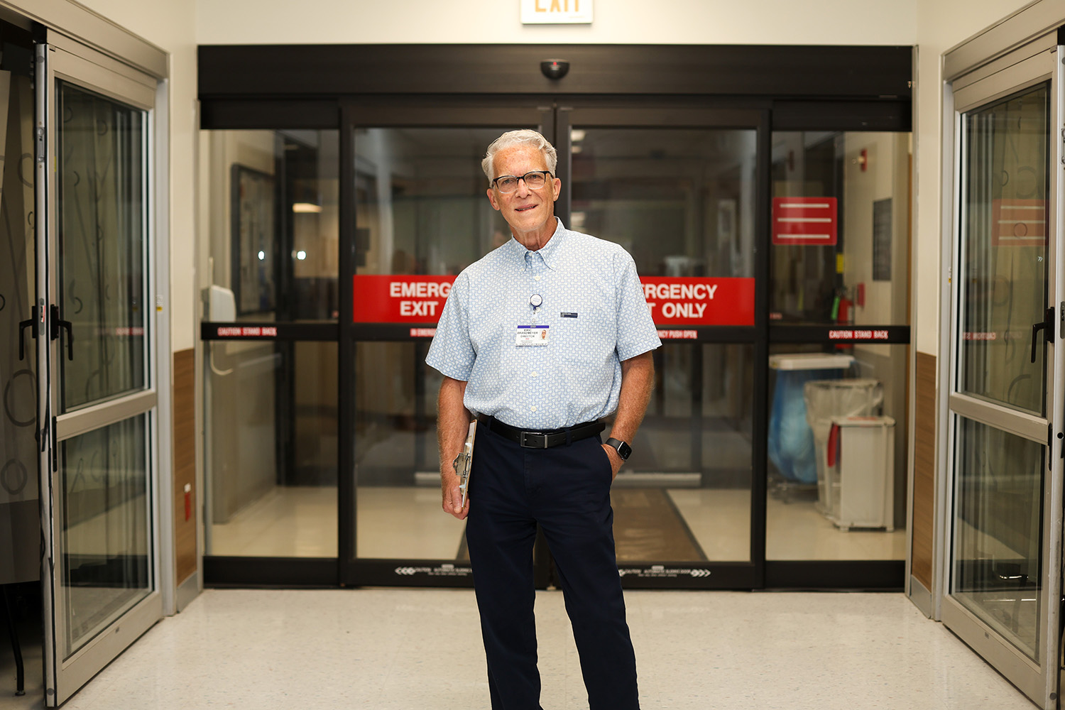 An older man wearing glasses, a light blue shirt, and dark pants stands smiling in a hospital hallway in front of glass doors labeled EMERGENCY EXIT ONLY. He has a staff badge clipped to his shirt.