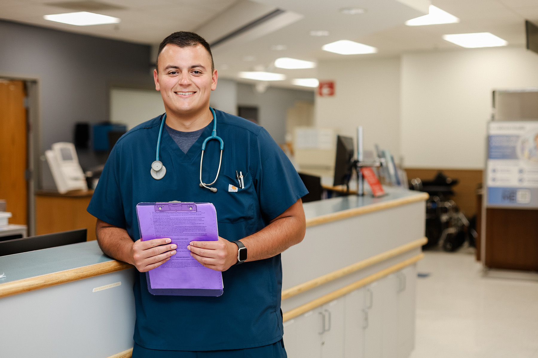A smiling healthcare worker in blue scrubs stands at a hospital counter, holding a purple clipboard. He wears a stethoscope around his neck and a smartwatch, with hospital equipment visible in the background.