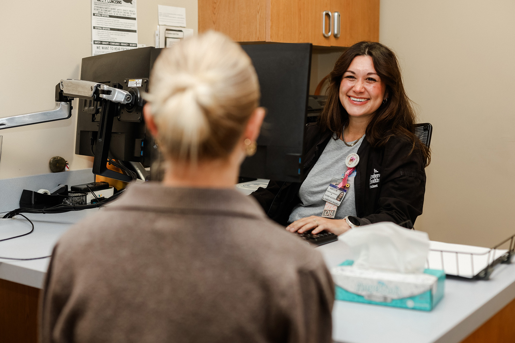 A smiling healthcare worker sits at a desk with a computer, talking to a patient whose back is to the camera. A box of tissues and papers are on the desk in a clinical office setting.