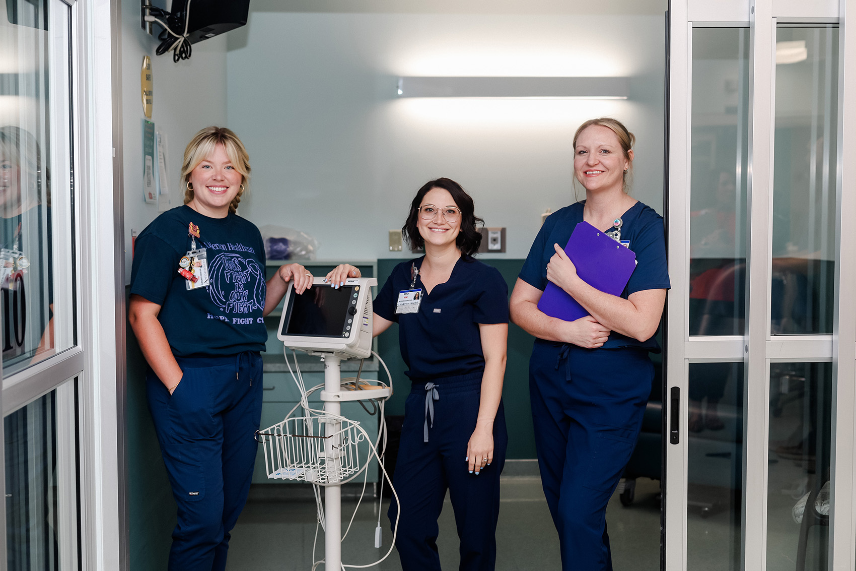 Three nurses in navy blue scrubs stand in a hospital room, smiling at the camera. One holds a clipboard, another stands by a medical monitor, and a third stands with hands in pockets. Medical equipment is visible around them.