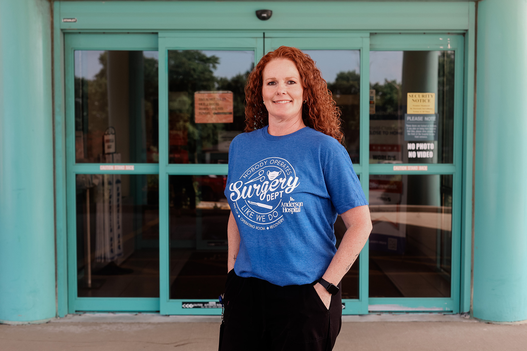 A woman with curly red hair stands smiling in front of glass doors. She is wearing a blue T-shirt that says “Surgery Open” and has one hand in her pocket. Signs on the doors read No Photo No Video.