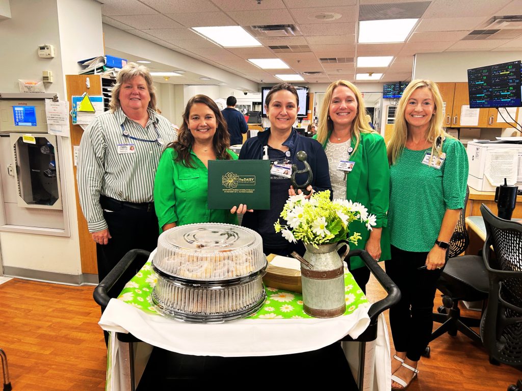 Five women stand behind a cart with flowers, baked goods, and a certificate in a brightly lit office or hospital setting, smiling at the camera.